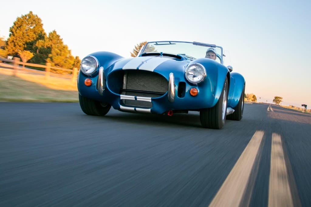 Blue Shelby Cobra replica with white racing stripes driving on an open road at golden hour
